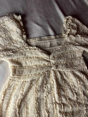 Blus  - Supersöt krämvit klänning med puffärmar och volangdetaljer över hela tyget. Smockad överdel och lätt rynkad kjol ger en romantisk vibe. Perfekt för sommarens alla tillfällen och har en mjuk, luftig känsla.