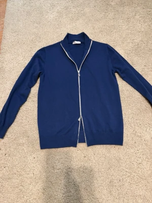 Blå kofta från Brunello Cucinelli - Säljer en otroligt mjuk och elegant marinblå full zip-cardigan från Brunello Cucinelli i cashmere. Klassisk design med ribbade detaljer, i väldigt bra skick – sparsamt använd. Nypris cirka 2800 euro. Inga kvitton kvar. Välkomnar seriösa prisförslag 