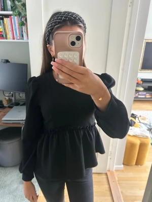 Svart blus med puffärmar - Säljer en svart blus med puffiga långärmar och markerad midja med volangkant. Blusen har rund halsringning och en lös, flowy passform nedtill. Perfekt att styla med jeans eller kjol för en trendig look.