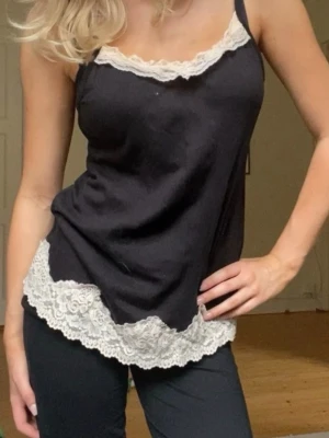 Svart linnetopp med spetskant - Svart linnetopp med tunna axelband och dekorativ vit spets längs både ringning och nederkant. Snygg och enkel modell som ger en feminin touch. Perfekt att styla med jeans eller kjol för en trendig look.