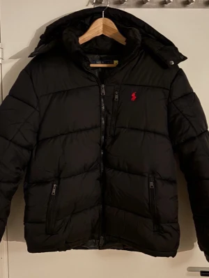 Svart pufferjacka från Polo Ralph Lauren - Säljer en svart pufferjacka från Polo Ralph Lauren med röd broderad logga på bröstet. Jackan har huva, dragkedja framtill och två sidofickor med dragkedja. Perfekt för kalla dagar och riktigt snygg till streetwear-stilen. Storlek S. Jättebra skick, endast provad. Pris kan alltid diskuteras så kom med bud. Hör av er vid frågor eller funderingar.