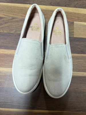 Beige mocka loafers från Dasia - Snygga beige loafers från Dasia i mjuk mocka. Skorna har elastiska paneler på sidorna för enkel påtagning och en vit platt sula. Perfekta för dig som gillar stilrena och bekväma skor till vardags. Använda 2 gånger