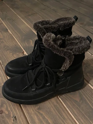 Svarta boots med fuskpäls, storlek 38 - Säljer ett par svarta boots med snörning och mjukt fuskpälsfoder i brunt. Skorna har en robust sula och är perfekta för kalla dagar. Utsidan är i mockaimitation och insidan är riktigt fluffig och varm. Passar dig som vill ha både stil och komfort under vintern. Mycket fint skick, knappt använda.