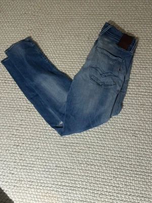 Blå replay anbass jeans med slitningar - Säljer ett par blå jeans från replay med slitningar på låret för en avslappnad look. Klassisk femficksmodell med ljus tvätt och skinnpatch bak i midjan. Perfekta till avslappnad eller uppklädd stil!