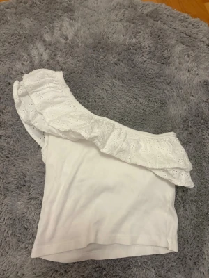 Vit oneshoulder topp med volang Gina Tricot - Supersöt vit oneshoulder topp från Gina Tricot med bred volangkant i broderad spets. Toppen är ribbad och har en ärmlös design med snygg asymmetrisk axel. Perfekt för sommarens alla tillfällen!