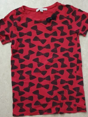 T-shirt - Röd t-shirt från Mywear Young med svarta rosetter över hela plagget och en extra tygrosett vid halsringningen. Klassisk rund hals och korta ärmar. Tillverkad i mjuk bomull som känns skön mot huden. Perfekt för dig som gillar lekfulla detaljer.