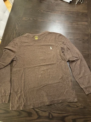 Dinozz tröjor - Säljer en brun långärmad tröja från Polo Ralph Lauren med klassisk rund hals och den ikoniska vita logon broderad på bröstet. Tillverkad i mjuk bomull som känns skön mot huden. Perfekt för dig som gillar enkel och stilren design.