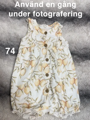Vit byxdress med citronmönster Newbie - Supersöt vit byxdress från Newbie med gult citronmönster och gröna blad. Byxdressen har volangdetaljer vid axlarna och knappar framtill. Perfekt för varma dagar och riktigt gullig stil.🌸Använd en gång under fotografering. Storlek 74  