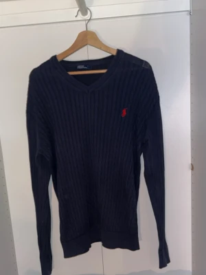 Mörkblå stickad tröja Ralph Lauren - Snygg mörkblå stickad tröja från Ralph Lauren med klassisk kabelstickning och röd broderad logga på bröstet. Tröjan har v-ringad hals och långa ärmar, perfekt för dig som gillar stilrena och tidlösa plagg.