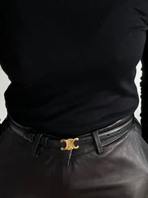 Svart Triomphe bälte i skinn - Säljer ett svart bälte med ikoniskt Triomphe-spänne i guld. Bältet är tillverkat i äkta skinn och har en stilren, klassisk form. Spännet är i metall med Celine Paris-gravyr. Perfekt för dig som gillar exklusiva accessoarer med statement-detaljer.