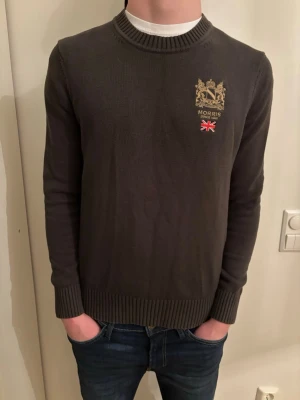 Mörkgrön stickad tröja från Morris - Snygg mörkgrön stickad tröja från Morris med broderad logga på bröstet. Rund hals och ribbade muddar vid ärmslut och nederkant. Perfekt för dig som behöver en varm tröja till vintern.