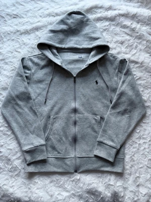 Grå zip hoodie från Polo Ralph Lauren - Snygg gråmelerad hoodie med dragkedja från Polo Ralph Lauren. Klassisk huva med dragsko, två fickor fram och ikonisk mörkblå broderad logga på bröstet. Mjuk bomullsblandning som är skön att ha på sig och passar perfekt till jeans eller joggers.