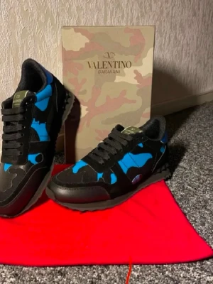 Valentino Garavani Rockrunner sneakers - Säljer ett par Valentino Garavani Rockrunner sneakers i svart och blått med coolt kamouflagemönster. Skorna har snörning, platt sula och detaljer i mocka och skinn. Perfekt för dig som vill sticka ut med en lyxig och urban stil.