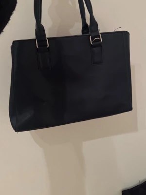 Svart axelväska i skinnimitation - Stilren svart axelväska med rejäla handtag och silverfärgade metalldetaljer. Väskan har en klassisk, fyrkantig form och är tillverkad i skinnimitation. Perfekt för dig som vill ha en enkel och snygg väska med plats för allt du behöver.