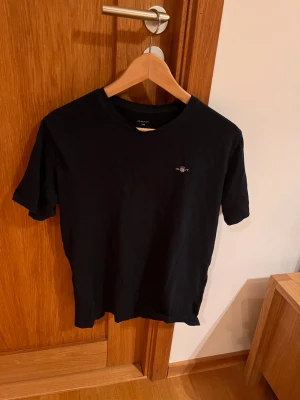 Svart t-shirt från GANT - Snygg svart t-shirt från GANT med liten broderad logga på bröstet. Klassisk rund hals och korta ärmar. Tillverkad i mjuk bomull som känns skön mot huden. Perfekt för en clean och stilren look. Hör av vid minsta fundering eller lägg ett prisförslag direkt😇