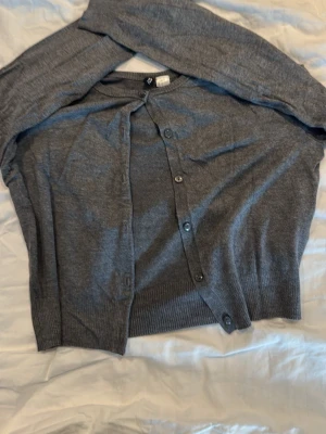 Grå kofta med knappar H&M - Mysig grå kofta från H&M med långa ärmar och knappar framtill. Klassisk modell med ribbade muddar vid ärmslut och nederkant. Perfekt att slänga över en t-shirt eller linne för extra lager.