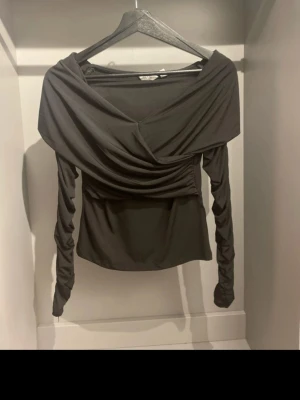 Svart omlottopp med draperade ärmar - Snygg svart omlottopp med bred, draperad urringning och långa, rynkade ärmar. Toppens material är mjukt och stretchigt, vilket ger en smickrande passform. Perfekt för dig som gillar en stilren men ändå unik look.