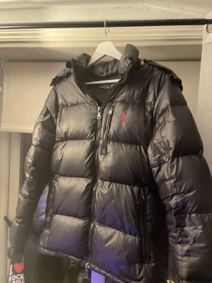 Svart pufferjacka från Polo Ralph Lauren - Säljer en svart pufferjacka från Polo Ralph Lauren med huva och dragkedja framtill. Jackan har en röd logga på bröstet och är riktigt fluffig och varm, perfekt för kalla dagar. Klassisk design med två sidofickor och quiltad look. DEN ÄR ÄKTA!!!