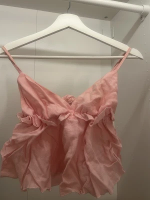 Ljusrosa volangig linnetopp - Supersöt ljusrosa linnetopp med tunna axelband och volangdetaljer framtill. Toppen har en luftig och flowy passform, perfekt för varma dagar. Materialet känns mjukt och lätt mot huden.