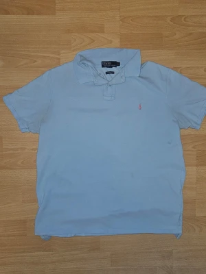 Ljusblå piké från Polo Ralph Lauren (M) - Ljusblå pikétröja från Polo Ralph Lauren i custom fit. Klassisk krage, korta ärmar och den ikoniska broderade loggan i rött på bröstet. Tillverkad i mjuk bomull som är skön att bära. Perfekt till jeans eller shorts för en clean look. Tag sizen stämmer inte riktigt. Den passar mer som en M eller stor S