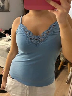 Ljusblå linnetopp med spets - Supersöt ljusblå linnetopp med tunna axelband och v-ringning dekorerad med spetsdetaljer framtill. Toppen är figurnära och gjord i mjukt material, perfekt för varma dagar eller att styla lager på lager.