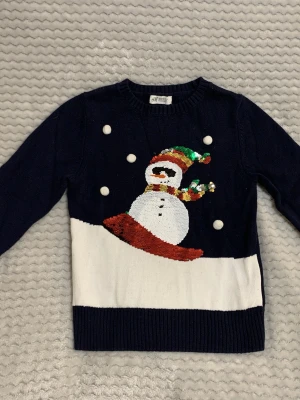Mörkblå jultröja med paljett-snögubbe H&M - Mysig mörkblå stickad tröja från H&M med en snögubbe i paljetter på framsidan. Tröjan har vita detaljer som snöbollar och en bred vit kant nedtill. Perfekt för jul och vinter, med långa ärmar och ribbade muddar.
