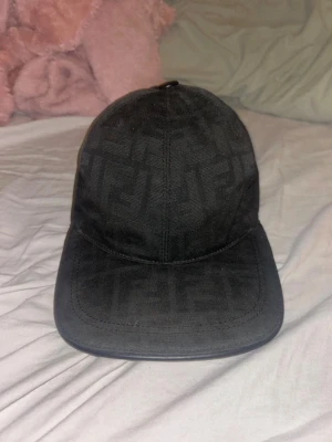 Svart Fendi keps med ton-i-ton mönster - Säljer en svart keps från Fendi med diskret ton-i-ton logomönster över hela kepsen. Justerbar rem bak i svart läder med Fendi-logga. Klassisk böjd skärm och stilren look som passar till det mesta.