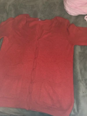 Röd kofta från Indiska - Säljer en röd kofta från Indiska i storlek M. Koftan har v-ringning, knappar framtill och två praktiska fickor. Perfekt för lager på lager och enkel att matcha med olika outfits. Materialet känns mjukt och skönt mot huden.