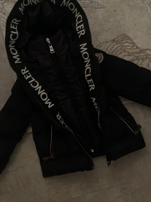 Svart Moncler pufferjacka med logga - Säljer en svart pufferjacka från Moncler med stor huva och tydlig logga längs kanten. Jackan har dragkedja framtill, två sidofickor med dragkedja och Moncler-märke på ärmen. Perfekt för kalla dagar och riktigt snygg streetstil.
