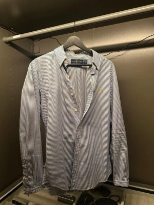 Blårandig skjorta från Ralph Lauren - Säljer en slim fit skjorta från Ralph Lauren i blått och vitt med klassiska vertikala ränder. Skjortan har lång ärm, knappar framtill och den ikoniska broderade loggan på bröstet. Tillverkad i mjuk bomull och perfekt för en clean och stilren look.