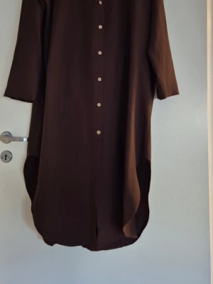 Set med long skorta och byxor - Brun skjortklänning med knappar framtill och lång ärm. Klänningen har en avslappnad passform och en stilren design.  Använd en gång.