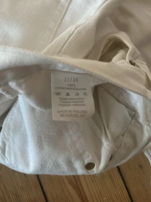 Vita raka jeans från Beavers - Säljer ett par klassiska vita jeans från Beavers i rak modell. Byxorna är tillverkade i 100% bomull och har fem fickor, metallnitar och en snygg etikett i midjan. Perfekta till en clean och stilren look.