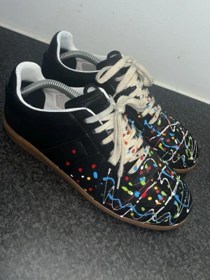 Maison Margiela sneakers med färgstänk - Ett par sköna Gats sneakers i svart skinn med unika färgstänk. Perfekt för dig som vill sticka ut med ett konstnärligt och edgy uttryck. Skorna är i nyskick, 9/10, och priset är satt därefter då kvitto o box saknas. Hör gärna av dig om du undrar över något!!! ‼️