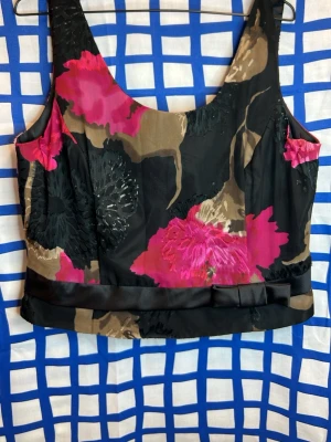 PLUS SIZE Blommig topp med rosett från Eurofashion - Snygg blommig topp från Eurofashion i svart, beige och stark rosa med breda axelband och dekorativ rosett i satin längst ner. Toppen har ett elastiskt smockparti i ryggen för skön passform och är tillverkad i ett glansigt, lätt material.