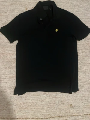 Svart pikétröja från Lyle & Scott - Klassisk svart pikétröja från Lyle & Scott med gul broderad logga på bröstet. Tröjan har korta ärmar, krage och knappar framtill. Tillverkad i mjukt bomullsmaterial som känns skönt mot huden. Perfekt till jeans eller shorts för en clean look.