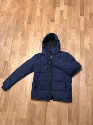 Mörkblå pufferjacka från Dressmann - Säljer en mörkblå pufferjacka från Dressmann i storlek M. Jackan har huva, dragkedja framtill och två sidofickor. Den quiltade designen ger en schysst look och jackan är perfekt för kyliga dagar. Materialet är syntet och jackan har långärmade ärmar.