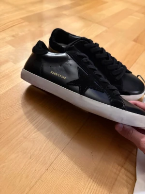 Golden Goose Super-Star svarta sneakers - Golden Goose Super-Star sneakers i svart skinn med vit sula och svart stjärndetalj på sidan. Klassisk låg modell med snörning och diskret guldtext på sidan. Kommer med dustbag. Perfekta för dig som gillar stilrena och exklusiva sneakers.