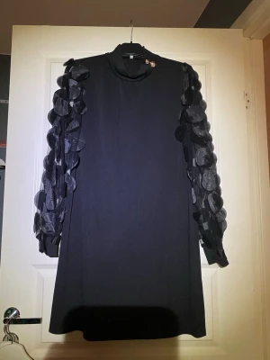 Svart kortklänning med meshärmar 2XL - Unik svart kortklänning med meshärmar dekorerade med stora applikationer i cirkelform. Hög krage med två guldknappar på axeln som snygg detalj. Klänningen har en rak passform och är perfekt för dig som vill sticka ut.