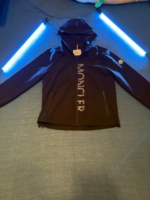 Svart vindjacka från Moncler - Helt ny !! Säljer en svart vindjacka från Moncler med huva och stora logotypen tryckt längs dragkedjan. Jackan har tre fickor med dragkedja och klassisk Moncler-patch på ärmen. Perfekt för dig som vill ha en clean och sportig look. Helt ny 