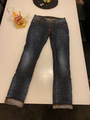 Mörkblå jeans från Nudie Jeans - Säljer ett par mörkblå jeans från Nudie Jeans med klassiska kontrastsömmar och snygga detaljer på bakfickorna. Jeansen har rak passform och är tillverkade i slitstarkt bomullsjeansmaterial. Perfekta för dig som gillar stilrena och tidlösa jeans.