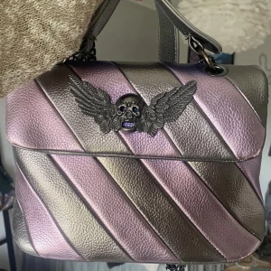 Maxjenny handväska - Säljer en snygg axelväska i metallic skimrande lila och silver med diagonala ränder. Väskan har ett coolt dödskalle med vingar i metall framtill. Perfekt statement piece för dig som gillar att sticka ut.  Längd: 26cm  Bredd: 8cm  Höjd: 20 cm