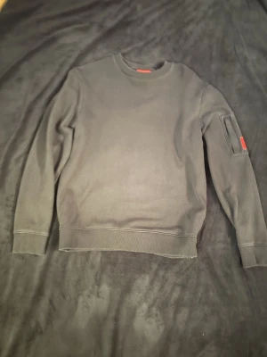 Svart hugo boss sweatshirt med ficka på ärmen - En stilren svart hugo boss sweatshirt med rund hals och ribbade muddar. Tröjan har en unik detalj med en ficka på ena ärmen och en liten röd etikett med Hugo på. Perfekt för dig som gillar minimalistisk design och vill ha något enkelt men ändå med en twist.