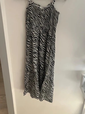 Zebramönstrad långklänning 152 - Säljer en långklänning med zebramönster i svart och vitt. Klänningen har smala, justerbara axelband och smock upptill för en bekväm passform. Perfekt för dig som vill sticka ut med ett djurmönster och gillar en luftig och skön stil.