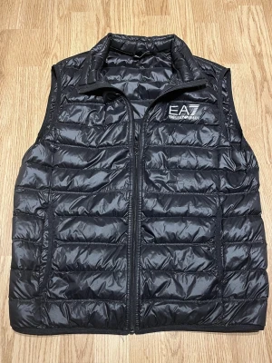 Svart väst från Emporio Armani EA7 - Säljer en svart dunväst från Emporio Armani EA7. Västen har hög krage och två sidofickor. Använd endast fåtal gånger och är i mycket fint skick utan defekter. Plagget har QR-kod för äkthetsverifiering. 