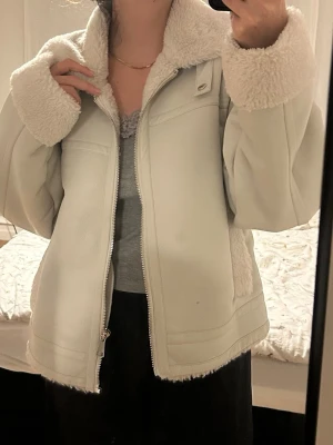 Vit teddyjacka med dragkedja - Säljer en supermjuk och fluffig vit teddyjacka med bred krage och muddar i fuskpäls. Jackan har en oversized passform och stängs med dragkedja framtill. Insidan är helfodrad med teddy och den har snygga detaljer längs kanterna. Perfekt för kalla dagar!