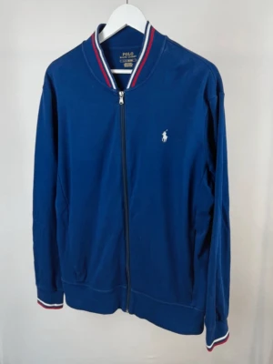 Blå zip-kofta från Polo Ralph Lauren - Snygg marinblå kofta från Polo Ralph Lauren med dragkedja framtill och klassisk vit logga på bröstet. Kragen och muddarna har röd och vit randig detalj. Tillverkad i mjuk bomull, perfekt för lager på lager under höst och vår.