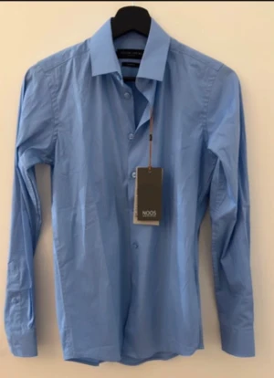 Blå slim fit skjorta från Casual Friday - Fräsch ljusblå skjorta från Casual Friday by Blend i slim fit-modell. Skjortan har klassisk krage, långa ärmar med manschett och knappar framtill. Tillverkad i mjuk bomull som känns skön mot huden. Perfekt för dig som gillar stilren och enkel look.