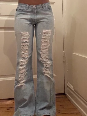 Ljusblå bootcut jeans med slitningar - Säljer ett par ljusblå jeans med rejält slitna detaljer framtill på båda benen. Byxorna har klassisk femficksmodell, hög midja och bootcut passform som ger en snygg retrovibe. Perfekta för dig som gillar en edgy och avslappnad stil.