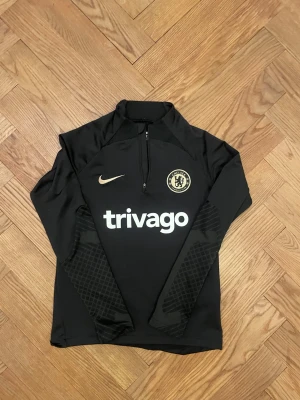 Chelsea Nike träningströja  - Svart Chelsea FC träningströja från Nike med guldiga detaljer och logga. Tröjan har halv dragkedja, Dri-Fit material och slim fit passform. Perfekt för fotbollsträning eller sportiga dagar. Snyggt mönster på ärmarna