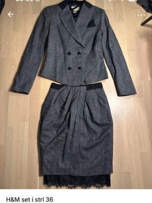 Grå randig kavaj och kjol från H&M - Säljer ett snyggt set från H&M i grått med diskreta ränder. Kavajen är dubbelknäppt med svarta detaljer vid krage och bröstficka. Kjolen har veck framtill, svarta detaljer i midjan och spetskant nedtill. Perfekt för dig som vill ha en stilren och modern look.